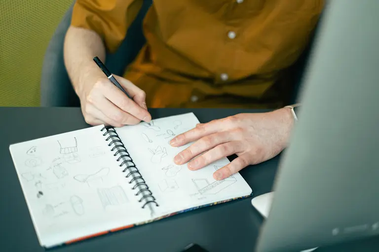 A designer sketching ideas in a spiral notebook beside a laptop