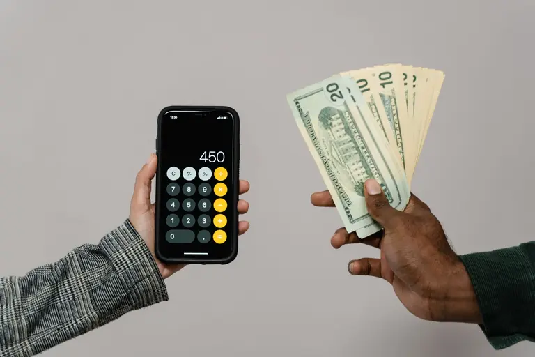 A hand holding a smartphone calculator while another hand offers a fan of banknotes