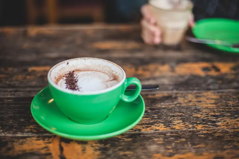 A cappuccino in a green cup on a rustic wooden table, representing the Halcyon Café brand system